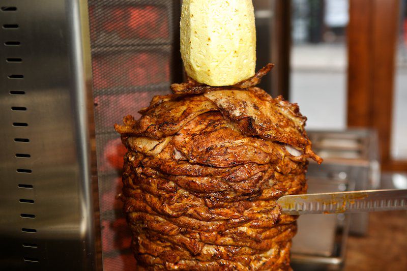 The Trompo turns with three sides being heated at a time. A skinned pineapple roasts above it, caramelizing along with the outer layer of the pork<br>